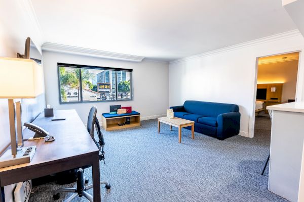 The image shows a bright, spacious room with a desk, chair, blue sofa, coffee table, and a large window displaying an outdoor view.