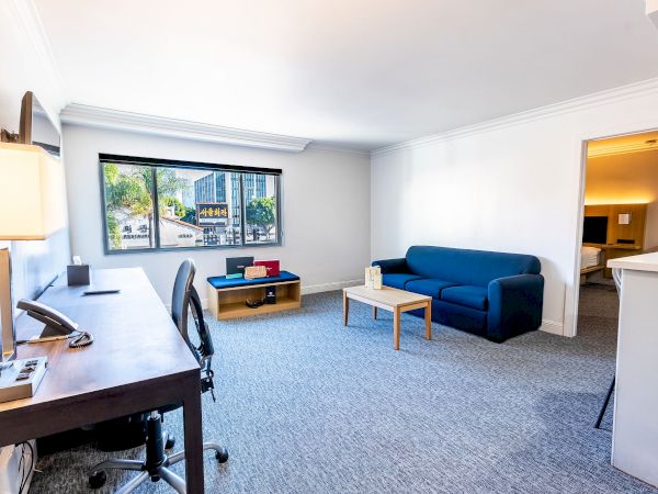 The image shows a bright, spacious room with a desk, chair, blue sofa, coffee table, and a large window displaying an outdoor view.