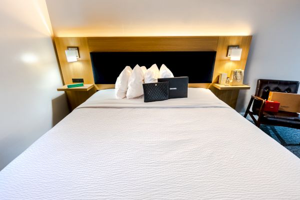 A neatly made bed with white linens, decorative pillows, a black box on top, and bedside tables with lamps and items, including a bottle and glasses.