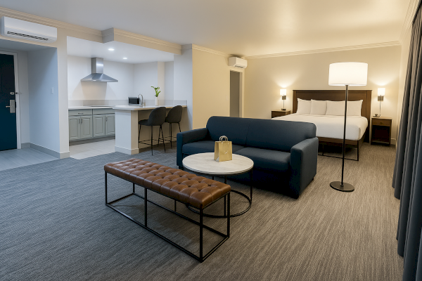 A modern hotel room with a bed, couch, table, bench, kitchenette, and floor lamp, featuring a spacious and comfortable layout.