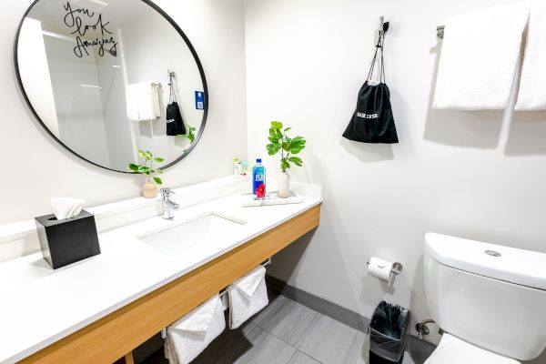 A modern bathroom with a round mirror, sink, plant, tissue box, hand soap, toilet, two towels, a black hanging bag, and a waste bin.