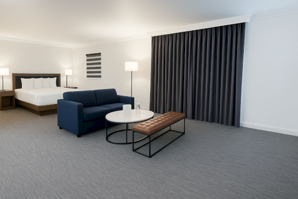 The image shows a modern hotel room with a bed, sofa, coffee table, bench, lamps, and dark curtains, creating a stylish, minimalist ambiance.