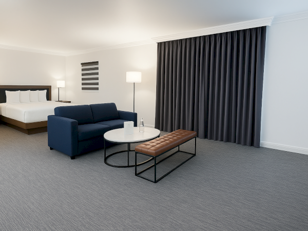 The image shows a modern hotel room with a bed, sofa, coffee table, bench, lamps, and dark curtains, creating a stylish, minimalist ambiance.