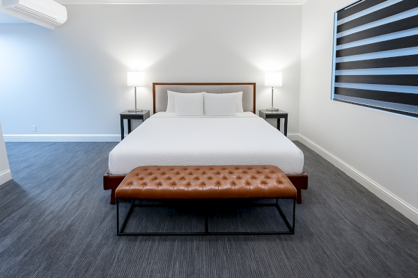A modern bedroom features a large bed with white bedding, two bedside tables with lamps, and a tufted bench at the foot.
