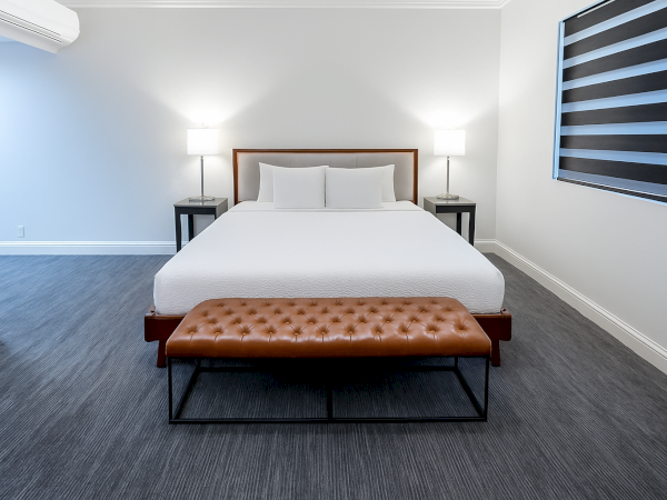 A modern bedroom features a large bed with white bedding, two bedside tables with lamps, and a tufted bench at the foot.