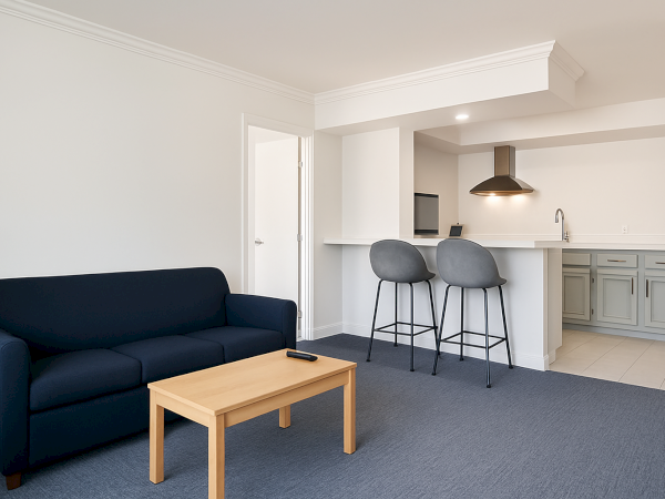 A modern living space with a navy sofa, wooden table, gray barstools, and an open kitchen layout featuring light cabinetry.