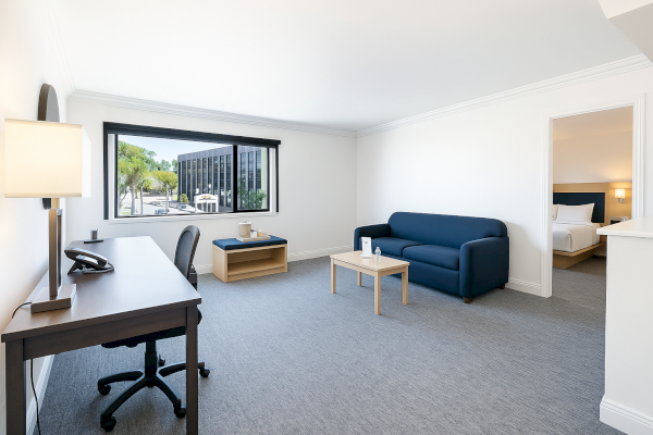 A modern, spacious room with a desk, chair, sofa, coffee table, and window view, adjacent to a bedroom.