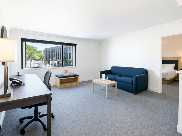A modern, spacious room with a desk, chair, sofa, coffee table, and window view, adjacent to a bedroom.