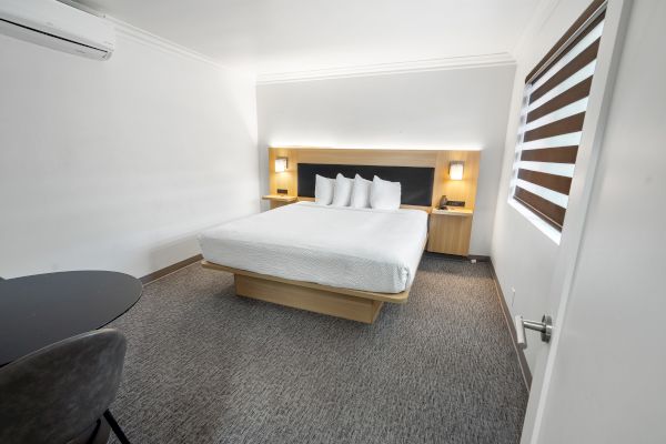 A minimalist hotel room with a large bed, two bedside tables with lamps, a chair and a table, and a wall-mounted air conditioner.