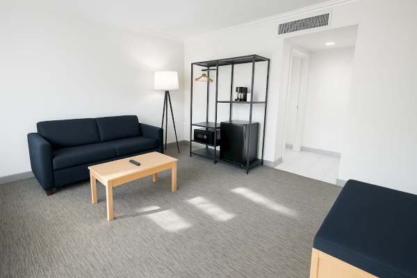 A modern living space with a blue sofa, lamp, coffee table, shelving, and a small fridge, in a minimalist style.
