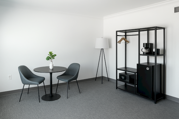 A minimalist room with a round table, two chairs, lamp, open shelving with hangers, coffee maker, safe, and mini fridge.