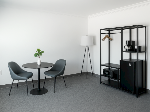 A minimalist room with a round table, two chairs, lamp, open shelving with hangers, coffee maker, safe, and mini fridge.