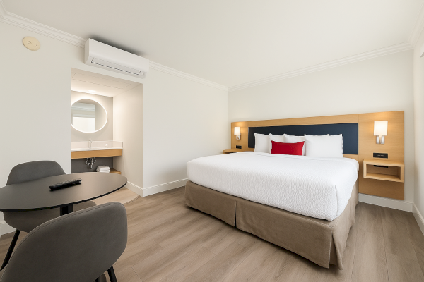 A minimalist hotel room with a large bed, table, two chairs, and a small vanity area with a circular mirror and sink.