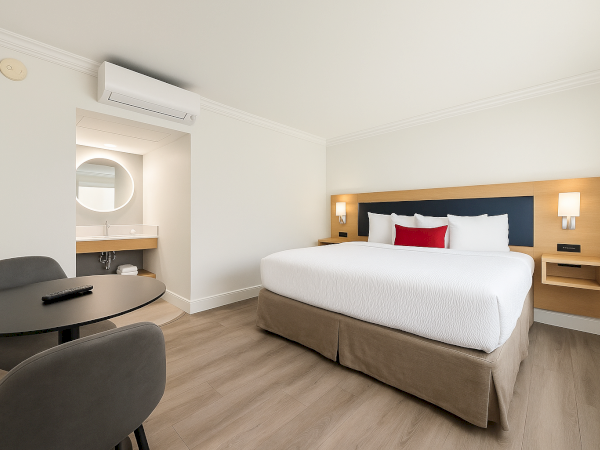 A minimalist hotel room with a large bed, table, two chairs, and a small vanity area with a circular mirror and sink.