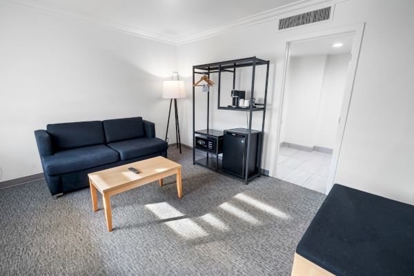 A minimalist living room with a black sofa, a wooden coffee table, a floor lamp, and a shelf holding a microwave and hangers.