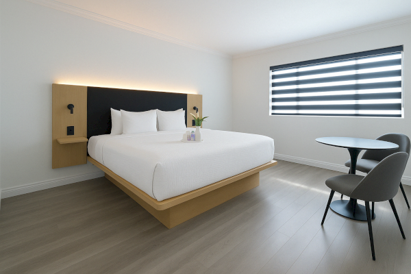 A modern bedroom with a minimalist design features a large bed, a table with two chairs, and a window with striped blinds.