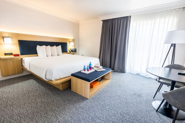 A modern hotel room with a large bed, bedside tables, white bedding, a bench, and a small table with chairs by the window.