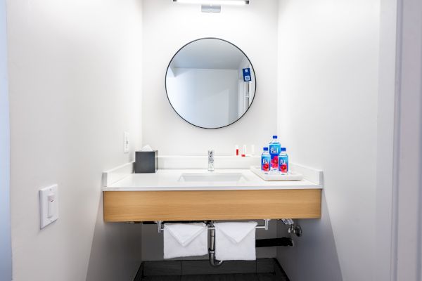 A minimalist bathroom features a round mirror, a sink, a soap dispenser, dental hygiene items, and two towels. Lighting is bright and clean.