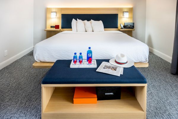 A modern hotel room with a neatly made bed, two water bottles, a white hat, and a newspaper on a bench; luxury boxes beneath.