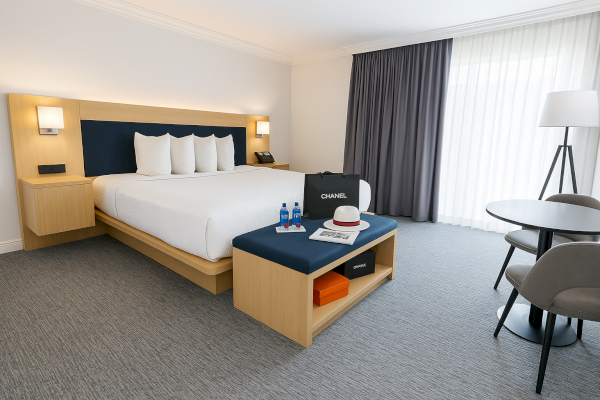 A modern hotel room with a large bed, side tables, a bag, and a hat on a bench, plus a table and chairs near the window.