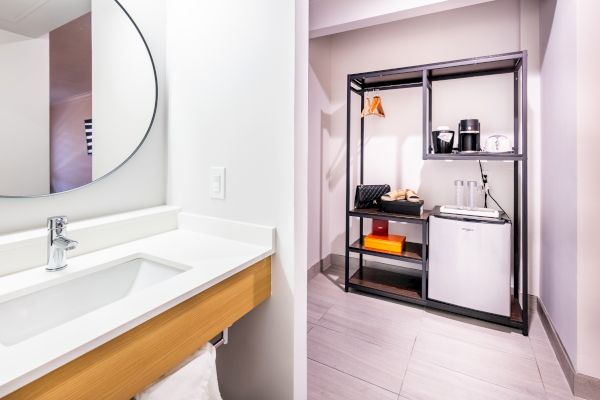 A modern bathroom with a sink and mirror is adjacent to a kitchenette area with a mini-fridge, microwave, coffee maker, and shelves for storage.