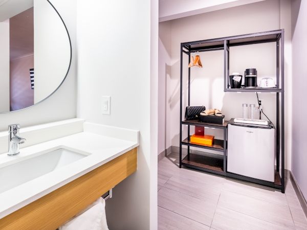 The image shows a modern bathroom with a sink and mirror on the left, and a kitchenette with a mini-fridge and coffee maker on the right.