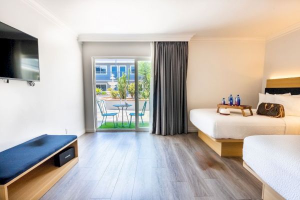 This image shows a bright hotel room with two beds, a wall-mounted TV, a small outdoor patio with seating, and a window with curtains.