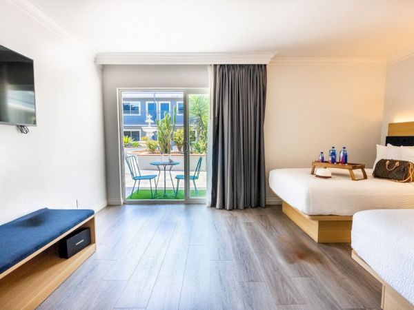 This image shows a bright hotel room with two beds, a wall-mounted TV, a small outdoor patio with seating, and a window with curtains.
