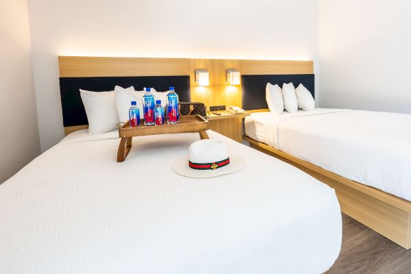 The image shows a modern hotel room with two beds, a tray of bottled water on one bed, and a white hat placed on the other bed, end of sentence.