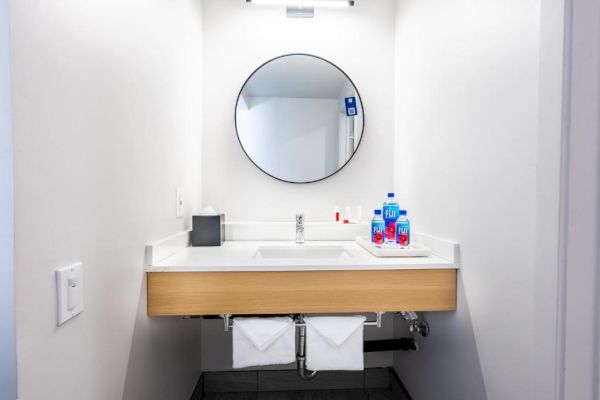 A bathroom sink with a round mirror, tissue box, hygiene products on the counter, towels below, and a modern, minimalist design.