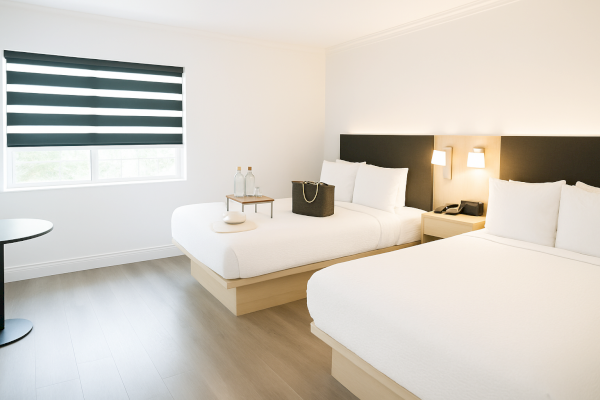 A minimalist hotel room with two beds, a window with striped blinds, and a small table holding two water bottles and a bag.
