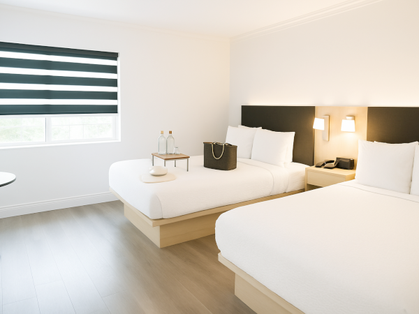 A minimalist hotel room with two beds, a window with striped blinds, and a small table holding two water bottles and a bag.
