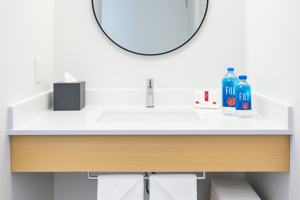 A minimalist bathroom vanity with a round mirror, tissue box, faucet, toiletries, two water bottles, and neatly folded towels below.