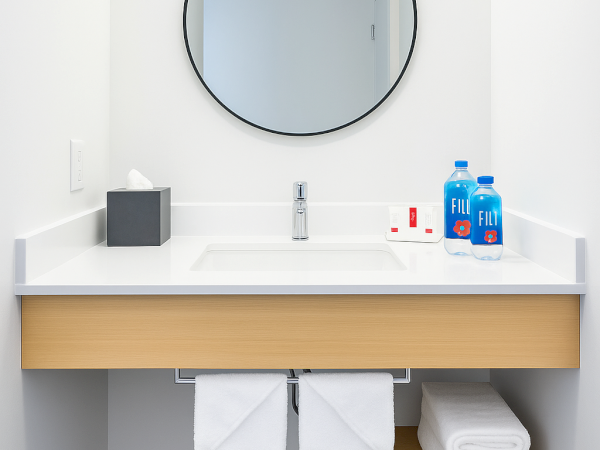 A minimalist bathroom vanity with a round mirror, tissue box, faucet, toiletries, two water bottles, and neatly folded towels below.