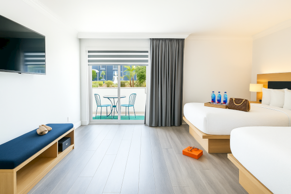 A hotel room with two beds, a TV, bench, a small table with chairs near a window, and items like bottles and a purse on the bed.