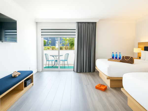 A hotel room with two beds, a TV, bench, a small table with chairs near a window, and items like bottles and a purse on the bed.