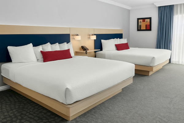 The image shows a hotel room with two beds, each with white bedding and red accent pillows. There’s modern lighting and framed art.