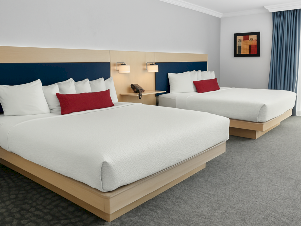 The image shows a hotel room with two beds, each with white bedding and red accent pillows. There’s modern lighting and framed art.