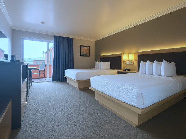 The image shows a hotel room with two large beds, a TV, and sliding glass doors leading to a balcony.