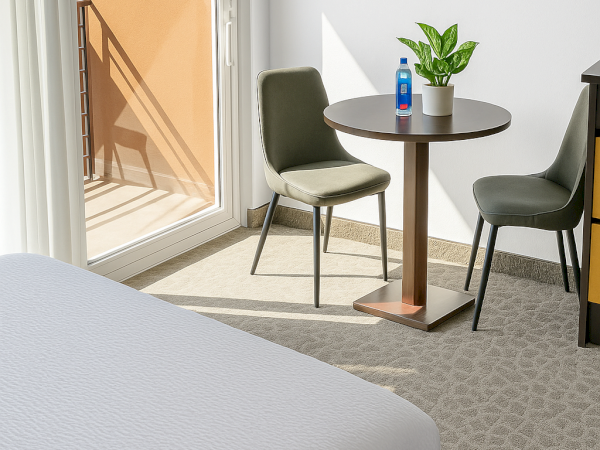 A modern room with a bed, a small round table with a plant, a water bottle, two chairs, and a sunny window with a balcony view.