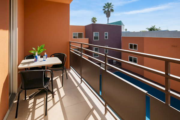A balcony with a table, two chairs, two water bottles, and a plant overlooks colorful buildings under a clear sky.