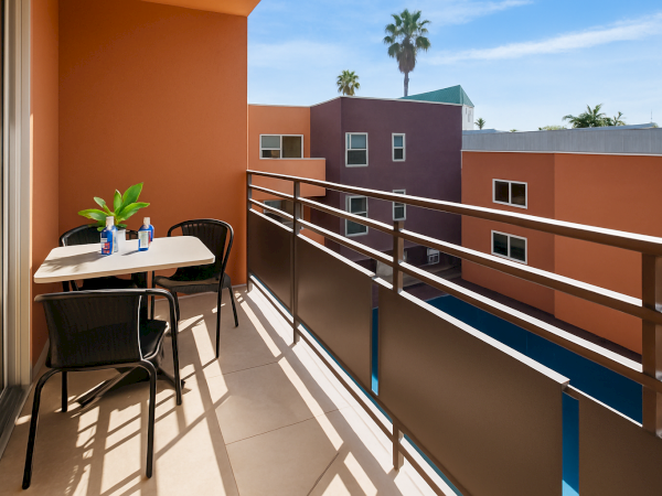 A balcony with a table, two chairs, two water bottles, and a plant overlooks colorful buildings under a clear sky.