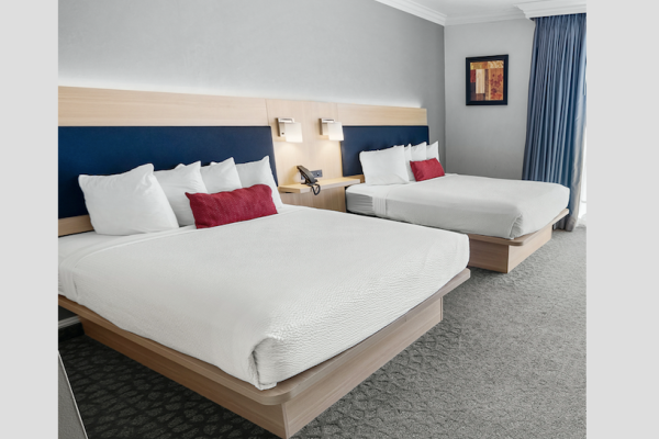 This image shows a hotel room with two made beds, white bedding, red and white pillows, wall lamps, a framed picture, and blue curtains.