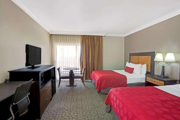 A hotel room with two double beds, red bedspreads, a TV, desk, chair, small table with two chairs, and a large window with curtains.