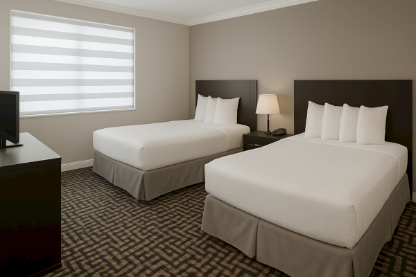 A hotel room with two neatly made beds, a nightstand with a lamp, a window with blinds, and a TV on a dresser in neutral tones.