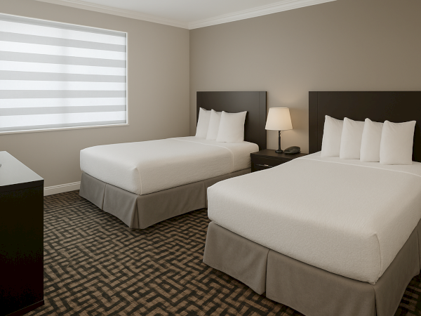 A hotel room with two neatly made beds, a nightstand with a lamp, a window with blinds, and a TV on a dresser in neutral tones.