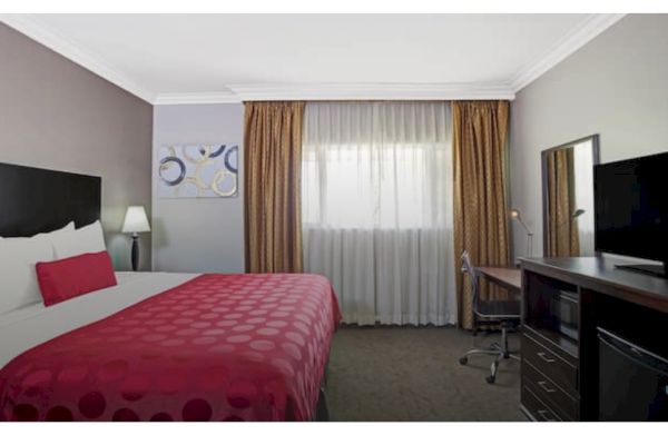 The image shows a hotel room with a bed, red bedding, a lamp, a window with curtains, a desk, chair, TV, and a wall art piece.