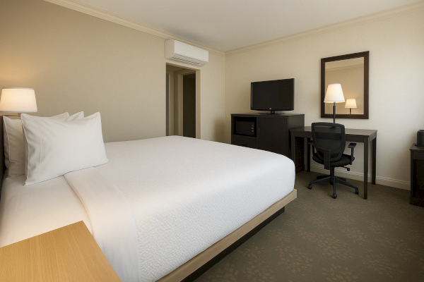 The image shows a modern hotel room with a large bed, desk, chair, TV, and lamp, decorated in neutral tones.