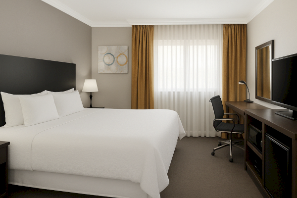 A hotel room with a bed, lamp, curtains, desk, chair, TV, and wall art.