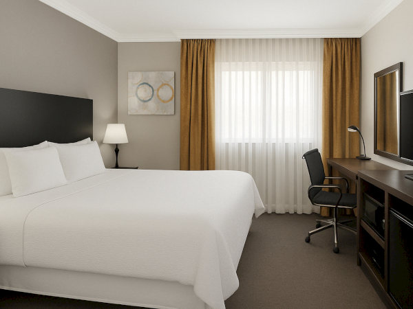 A hotel room with a bed, lamp, curtains, desk, chair, TV, and wall art.